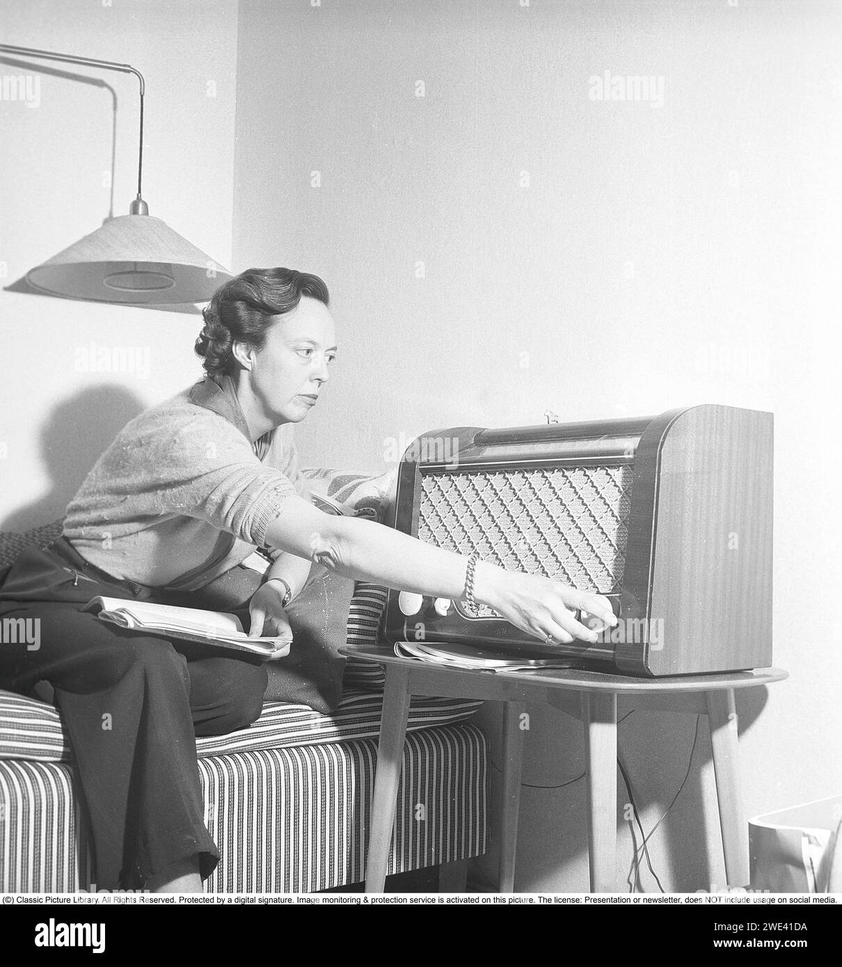 Ingrid Samuelsson, schwedische Radiojournalistin, war eine der ersten weiblichen Radioreporterinnen im schwedischen Radio, das vor den 1940er Jahren vollständig von Männern dominiert wurde. Hier zu Hause von einem Radio der Marke AGA. Sie dreht den Knopf, um die richtige Radiofrequenz für den Empfang der Radiosendung einzustellen. Der obere Teil des Radios zeigt an, welcher Sender gesendet wird, wenn Sie das Wahlrad drehen. Neben UKW-Sendungen wurden auch ausländische Radiosender auf Langwelle gehört. 1950 Krisoffersson Ref. AY3-4 Stockfoto