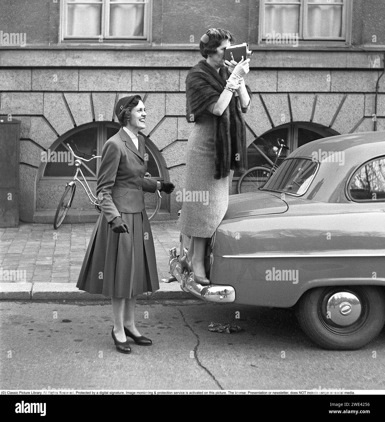 In den 1950er Jahren Eine ältere Frau mit einer Filmkamera stand auf dem Stoßfänger eines Autos, um einen besseren Blick auf das zu bekommen, was sie gerade filmt. Eine jüngere Frau steht daneben und sieht glücklich aus. Sie sind Verwandte eines der kürzlich abgeschlossenen Studenten, die feiern, dass sie ihre Immatrikulationsprüfung bestanden haben und eine Stockholmer Straße hinunterlaufen. 1954. Svahn Ref. SVA2 Stockfoto