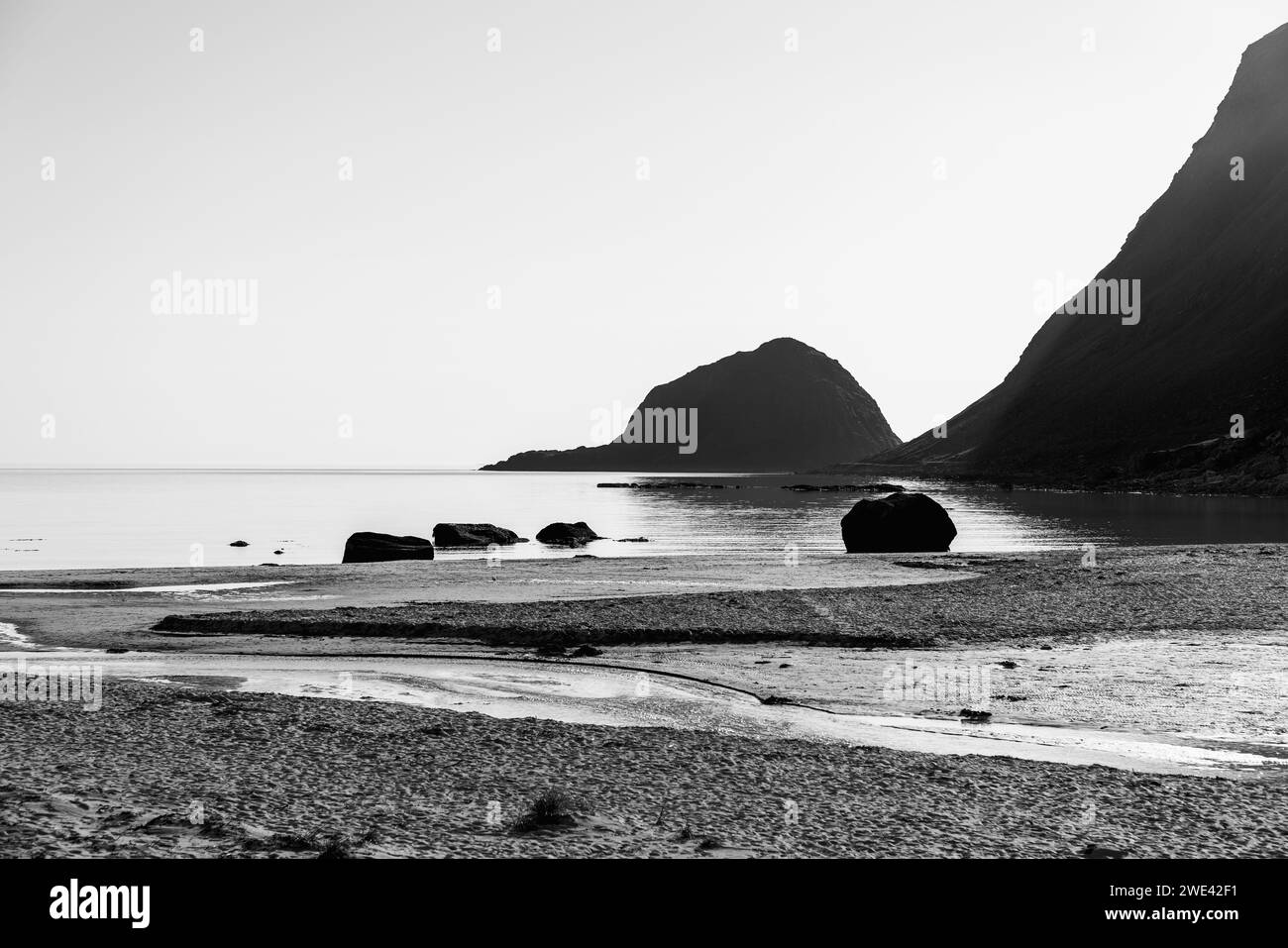 Starke Schwarz- und Weißtöne unterstreichen die ruhige Schönheit der Küste von Haukland Beach mit verstreuten Felsen und einer Bergsilhouette. Lofoten, Norwegen Stockfoto