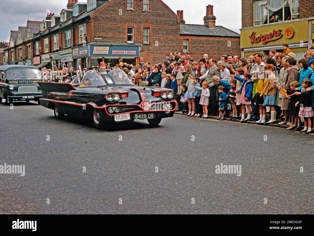 Watfords jährliche Pfsun-Karnevalsparade entlang der St Albans Road, Watford, Hertfordshire, England, Vereinigtes Königreich, 1967. ein Nachbau von Batmobile nahm Teil – das Auto erschien 1939 im DC-Comic Batman und in TV-Serien der späten 1960er Jahre – es zeigte Batman (Bruce Wayne) und sein Freund Robin (ein maskierter Robin befindet sich auf dem Beifahrersitz) benutzte ihr Fahrzeug, um Verbrechen zu verhindern und Bösewichte zu fangen. Ein Watford Observer Bedford Van folgt dem Batmobil – einem Vintage-Foto aus den 1960er Jahren. Stockfoto