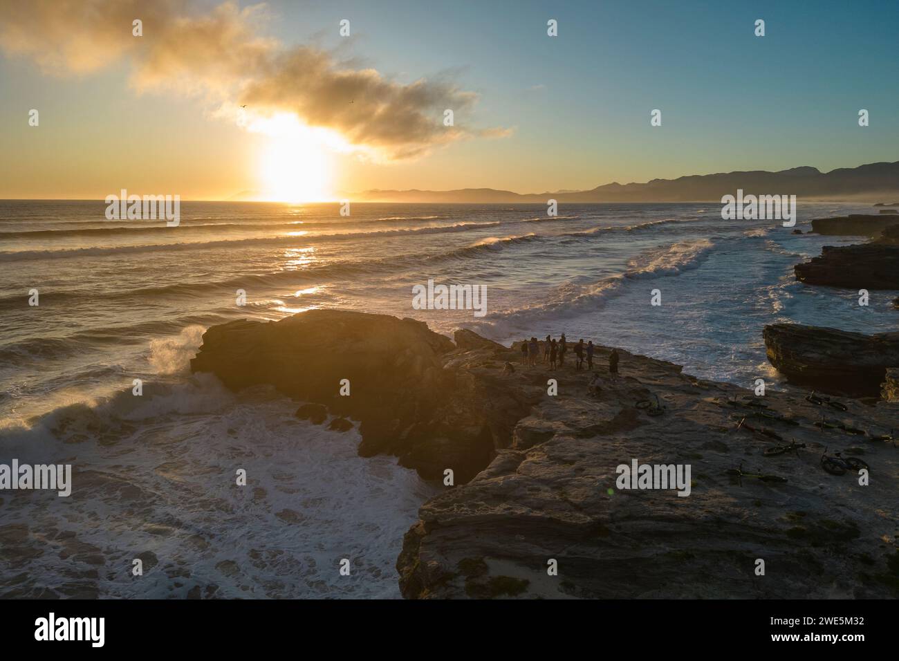 Aus der Vogelperspektive sehen Sie Menschen, die auf einem Felsvorsprung mit fetten Fahrrädern stehen, während Wellen bei Sonnenuntergang auf die Küste und den Strand im Walker Bay Nature Reserv brechen Stockfoto
