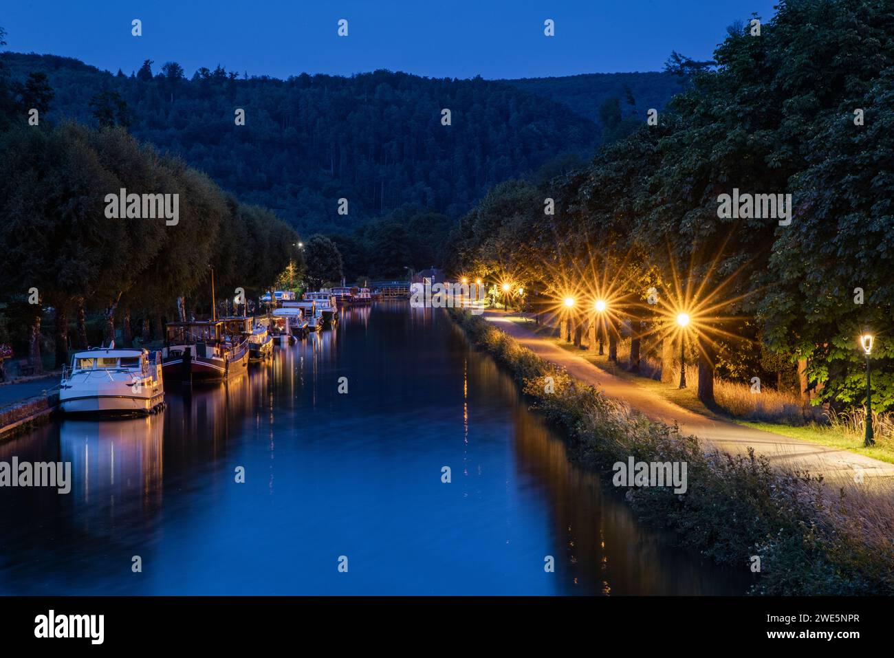 Hausboote, die auf dem Canal de la Marne au Rhin mit der Stadt in der Abenddämmerung, Lutzelbourg, Mosel, Frankreich, ankern Stockfoto