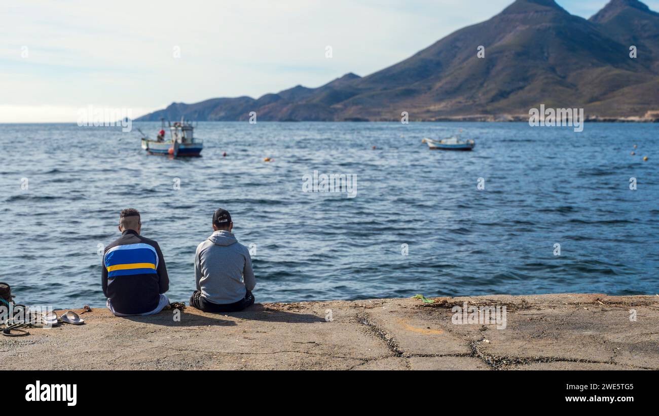 Zwei eingewanderte Jungs, die auf einem Pier an der Küste von Cabo de Gata, Almeria, auf das Meer schauen. Stockfoto