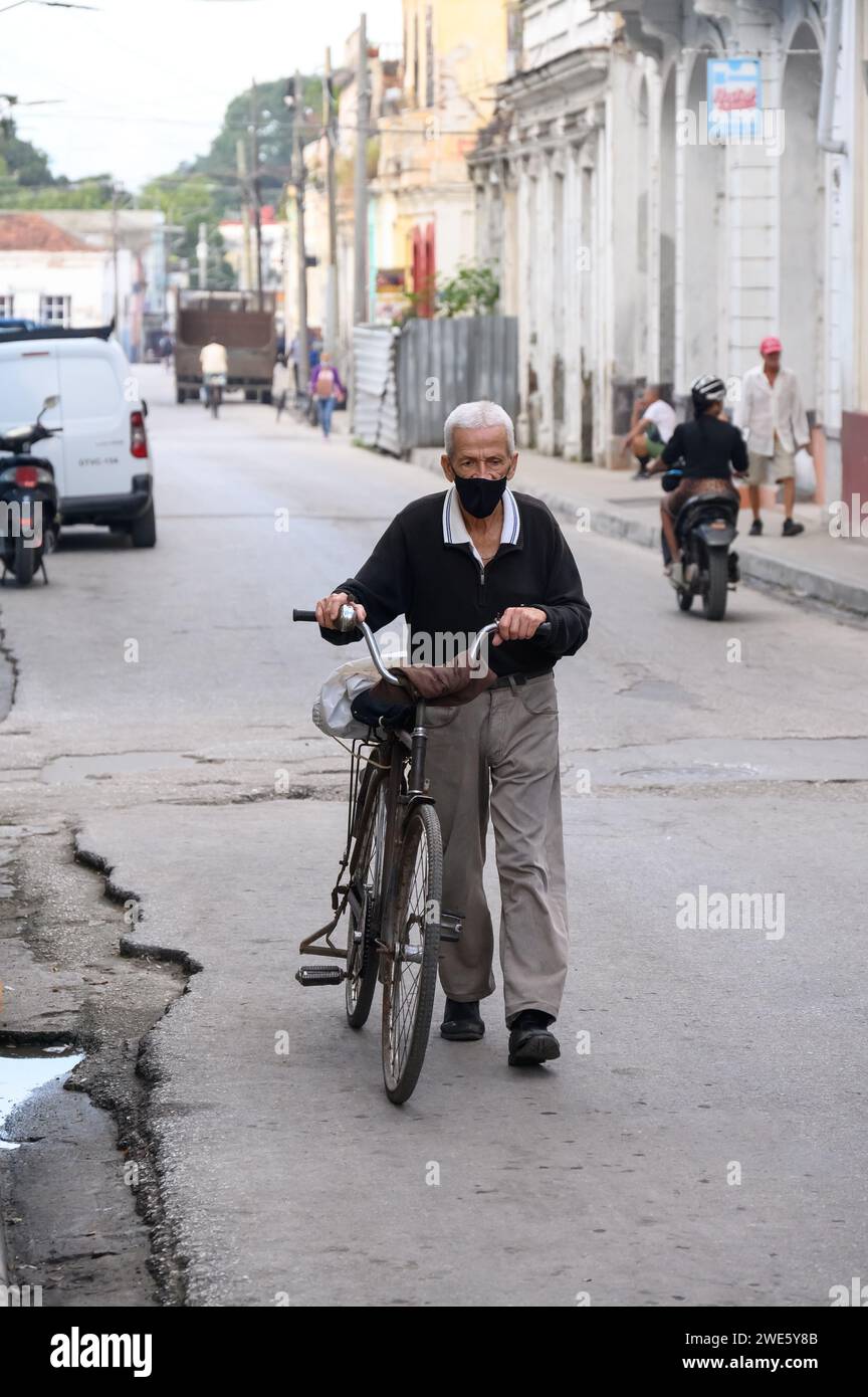 Ein älterer Mann mit Gesichtsmaske schiebt ein Fahrrad durch schwarzes Wasser in den Straßenrand. Stockfoto