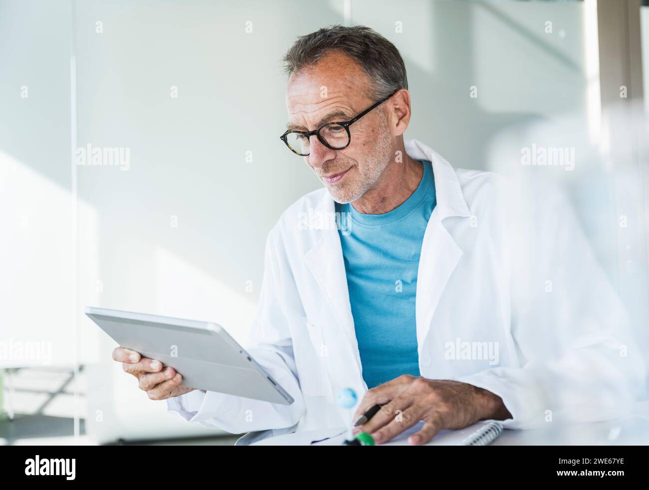 Lächelnder Arzt mit Tablet-PC und Notizblock am Schreibtisch Stockfoto