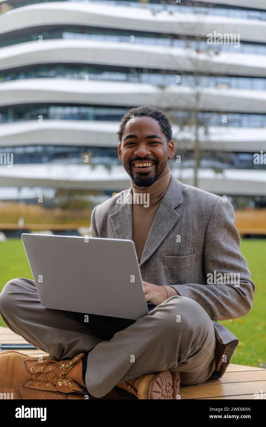 Fröhlicher Geschäftsmann, der mit Laptop auf der Bank im Büropark sitzt Stockfoto