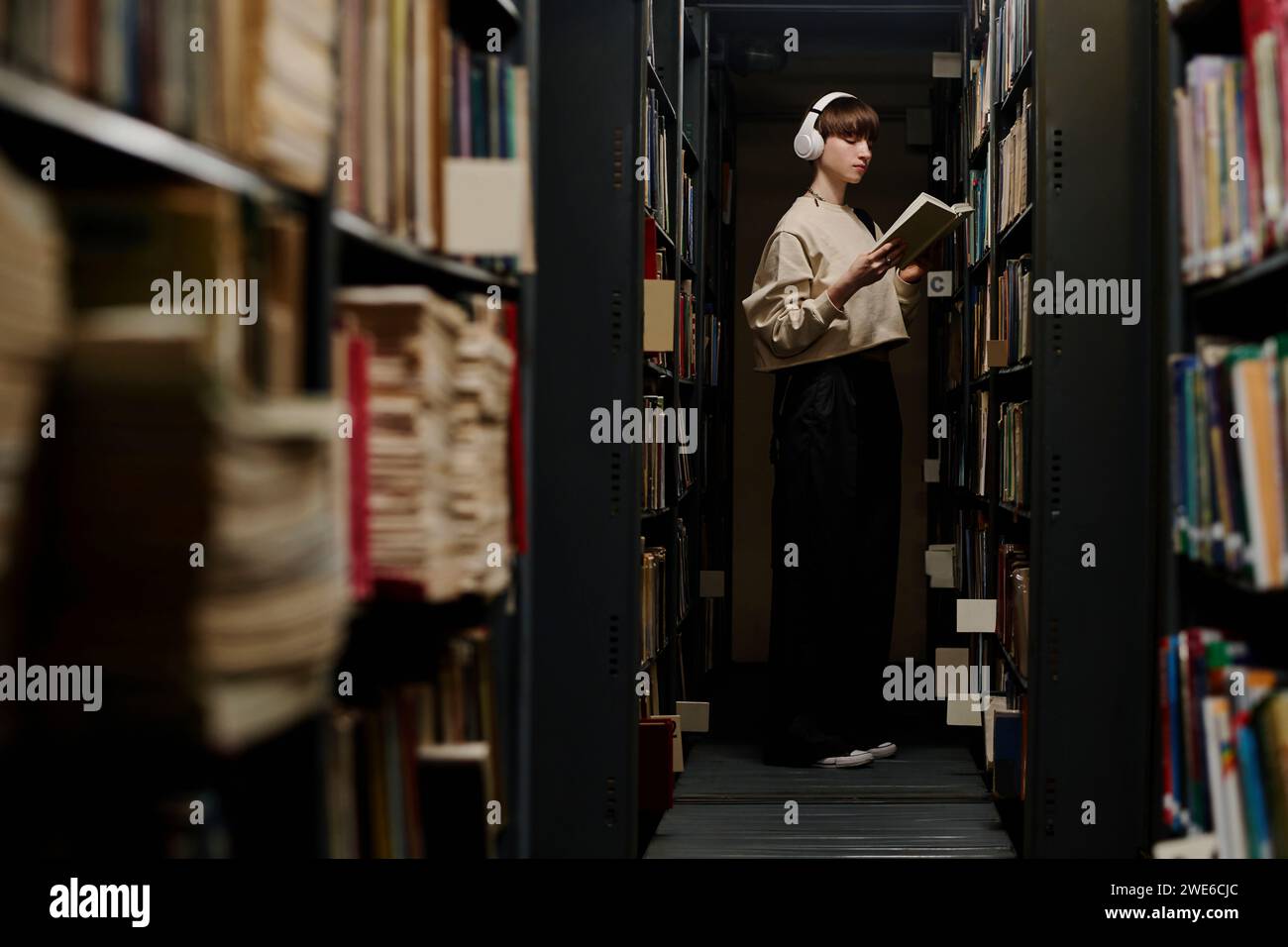 Der Schüler trägt kabellose Kopfhörer und liest Bücher in der Nähe der Regale in der Bibliothek Stockfoto