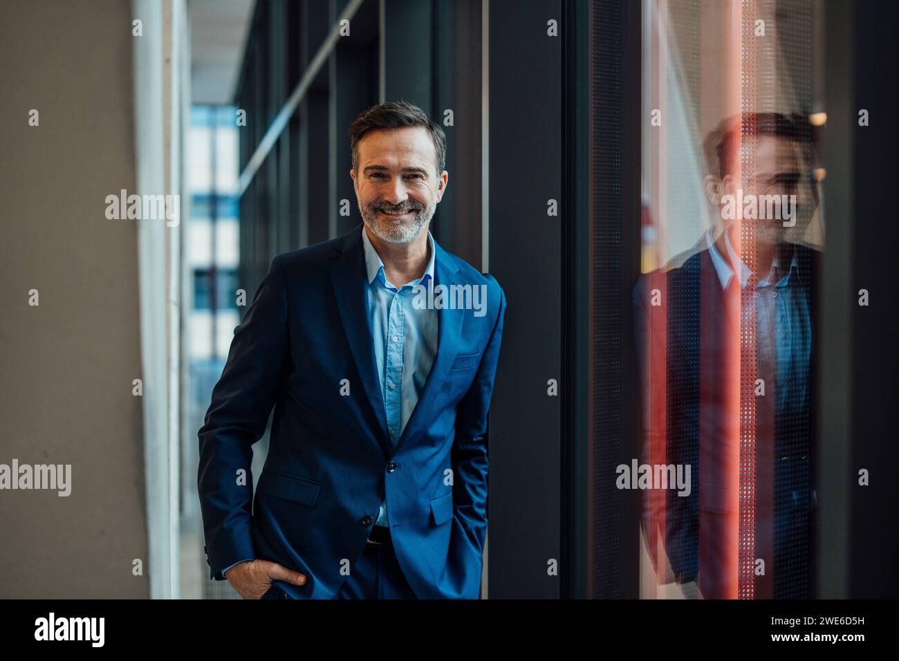 Lächelnder Geschäftsmann, der mit der Hand in der Tasche in der Nähe des Fensters steht Stockfoto