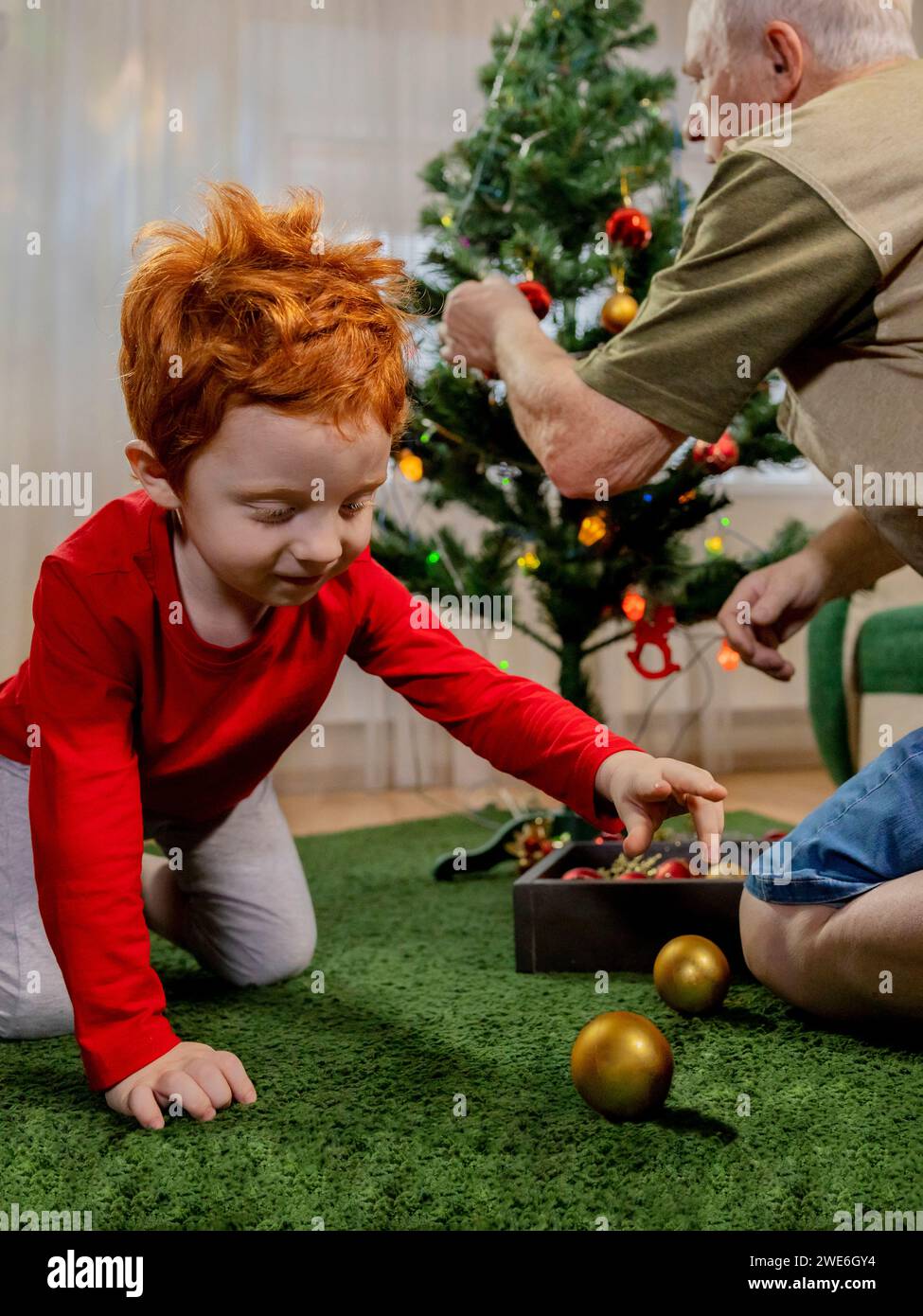 Rothaariger Junge, der mit der Kugel spielt, in der Nähe von Großvater, der zu Hause den Weihnachtsbaum dekoriert Stockfoto