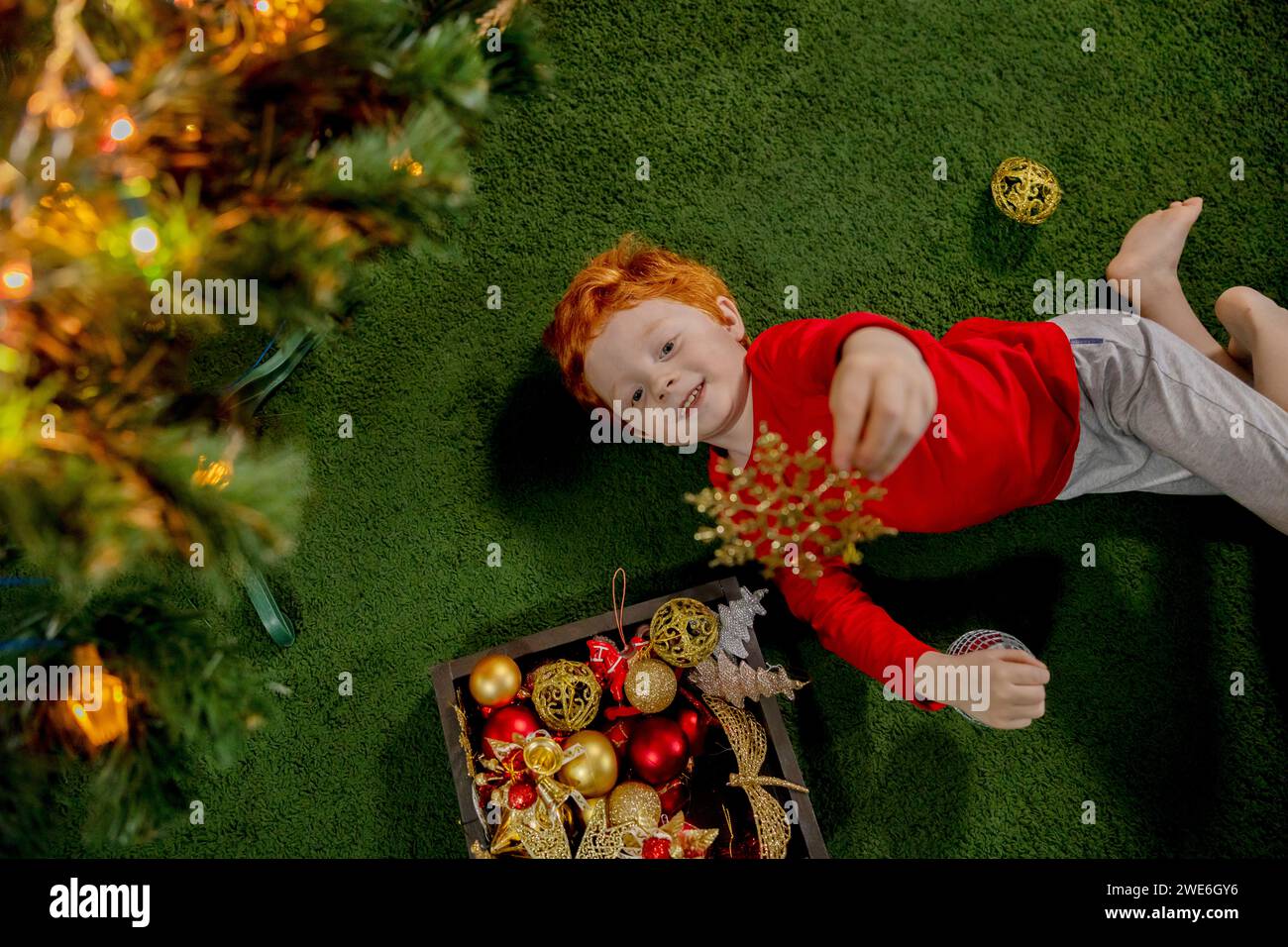 Glücklicher rothaariger Junge, der mit Weihnachtsschmuck auf grünem Teppich spielt Stockfoto Glücklicher rothaariger Junge, der mit Weihnachtsschmuck auf grünem Teppich spielt Stockfoto