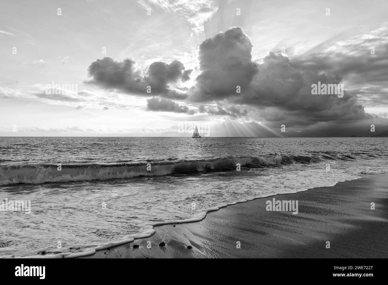 Ein Segelboot segelt entlang des Ozeans mit Sonnenstrahlen, die durch die Wolken in Schwarz und weiß brechen Stockfoto