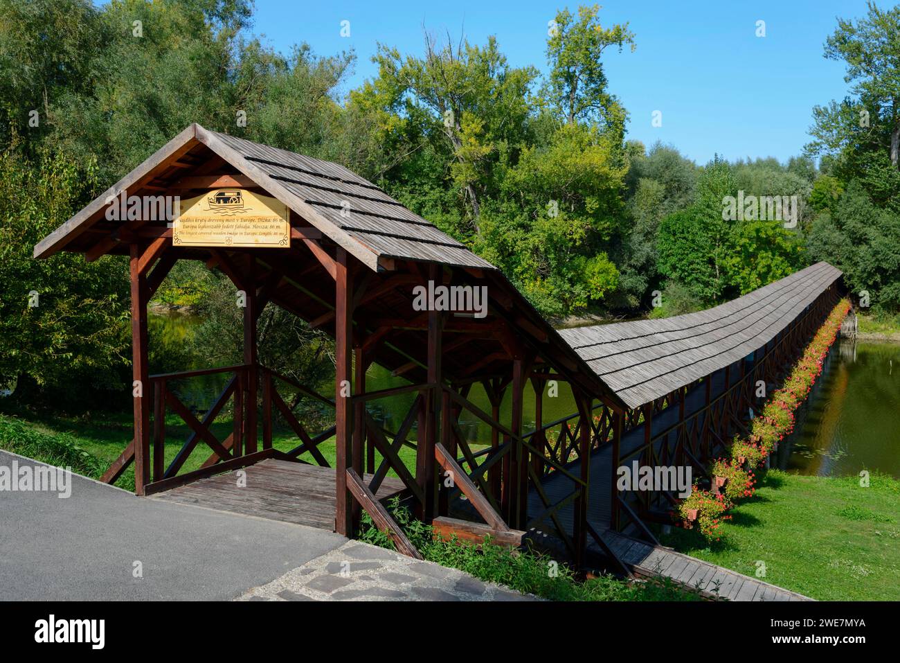 Eine überdachte Holzbrücke überspannt einen ruhigen Fluss an einem sonnigen Tag mit einem klaren blauen Himmel an der Schiffsmühle Kolarovo, Kolarovo, Guta, Komarno, Komarno Stockfoto