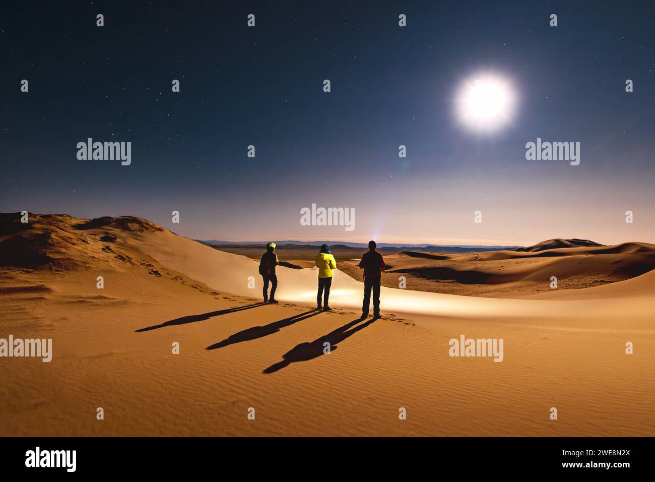 Drei Silhouetten von Wanderern mit Scheinwerfern stehen in der nächtlichen Wüstendüne und blicken auf den Sternenhimmel und den Mond Stockfoto