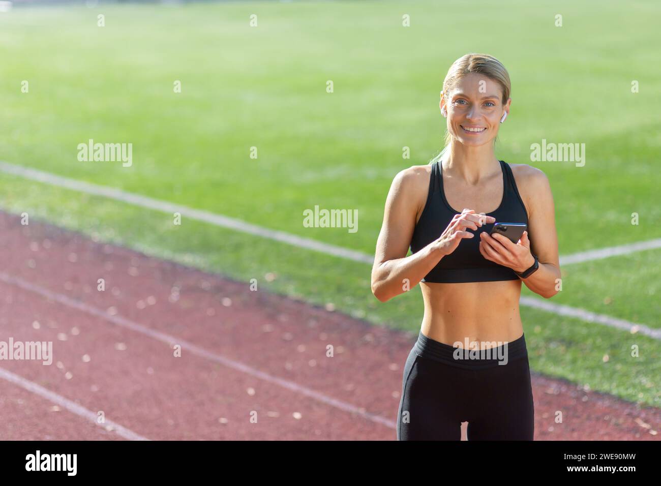 Eine lächelnde junge Frau in Fitnesskleidung steht auf einem Lauffeld, hält ein Telefon und bereitet sich auf ein Training vor. Stockfoto