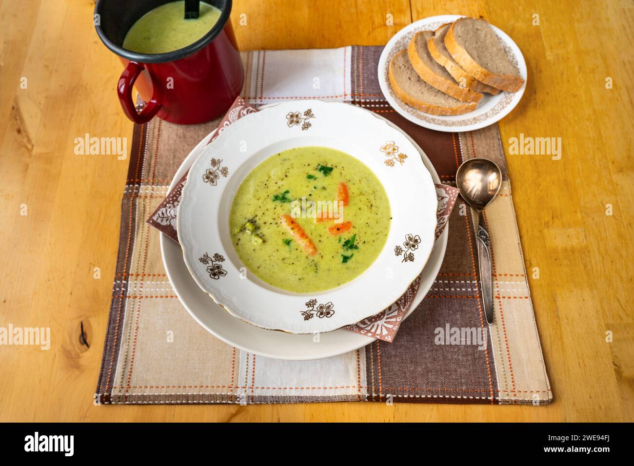 Cremige Brokkoli-Suppe im Teller und im Topf, Kelle, geschnittenes Brot auf Teller auf kariertem Tischtuch auf Holztisch. Stockfoto