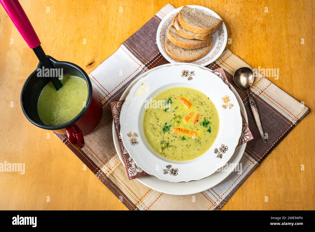 Cremige Brokkoli-Suppe in Teller und Topf, Kelle, Brotscheiben auf Teller auf Tischdecke auf Holztisch. Stockfoto