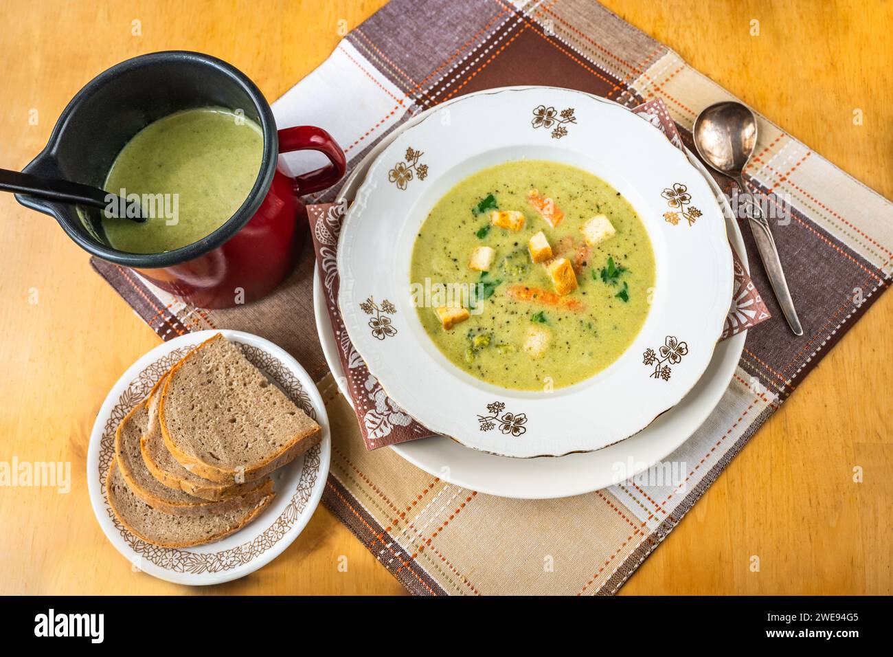 Cremige Brokkoli-Suppe in Teller und Topf, Brotscheiben auf Tischdecke auf Holztisch. Stockfoto