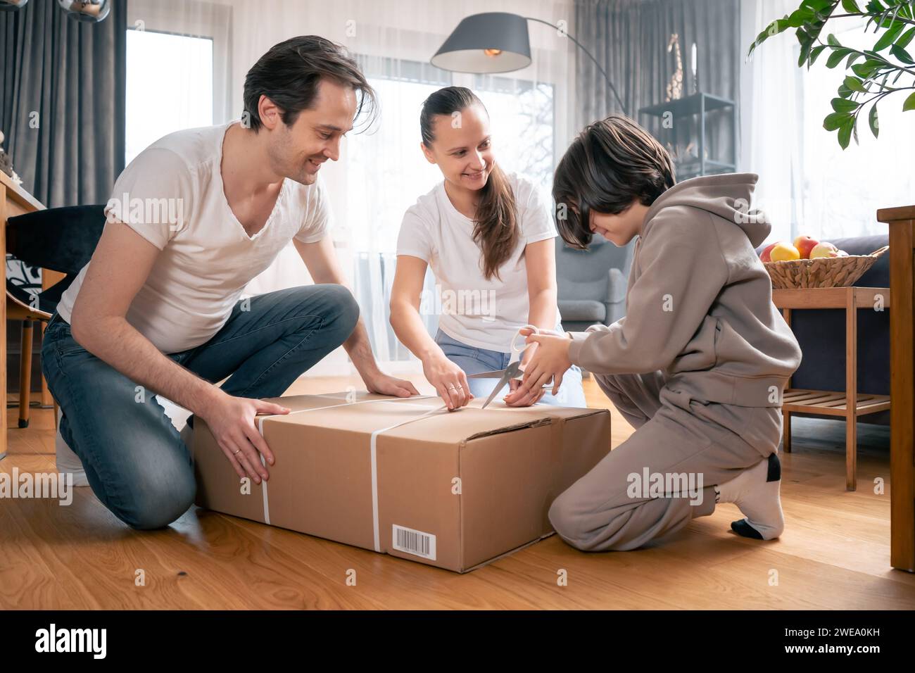 Sohn, Vater und Mutter packen gemeinsam einen großen Karton aus einem Paket mit Waren aus, die zu ihnen nach Hause geliefert wurden. Stockfoto
