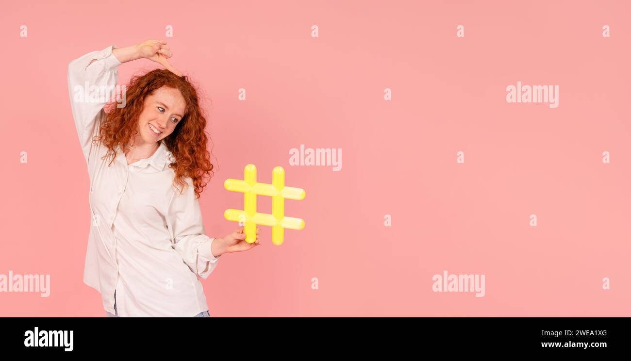 Aufgeregte Frau in weißem Hemd mit gelbem Hashtag in der Hand auf rosa Hintergrund mit Kopierraum. Soziale Medien, Blogging und virale Themen auf Stockfoto
