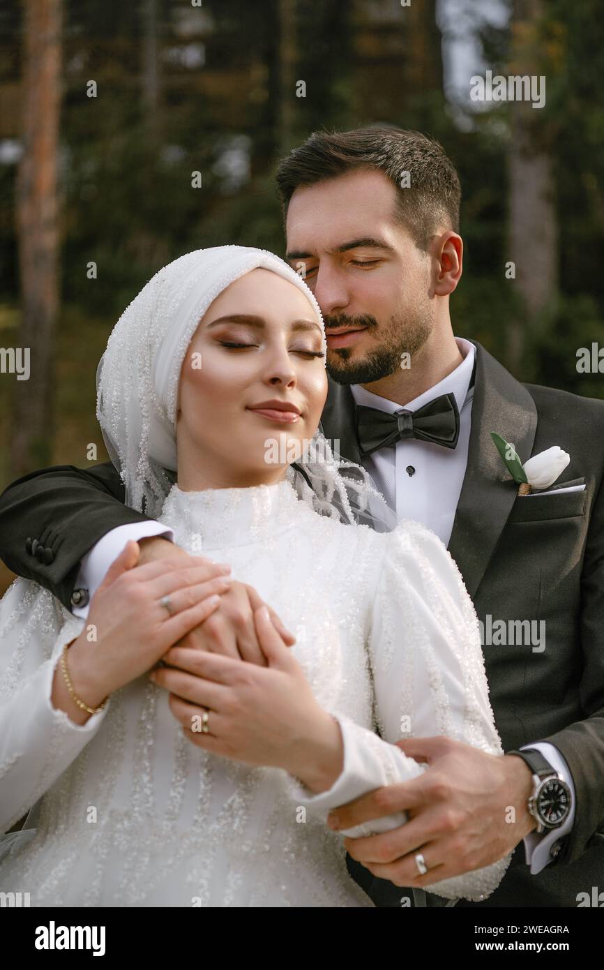 Der junge Bräutigam hat die Taille seiner Braut gewickelt und posiert mit geschlossenen Augen. Frisch vermählte Braut und Bräutigam feiern Hochzeit im Waldpark Stockfoto