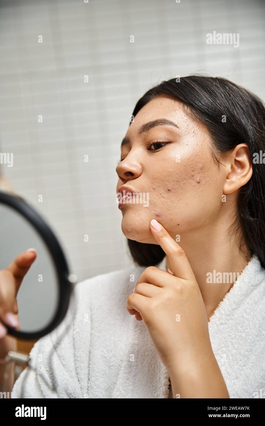 Junge asiatische Frau mit brünetten Haaren und Pickel, die ihr Gesicht im Spiegel untersucht, Hautprobleme Stockfoto