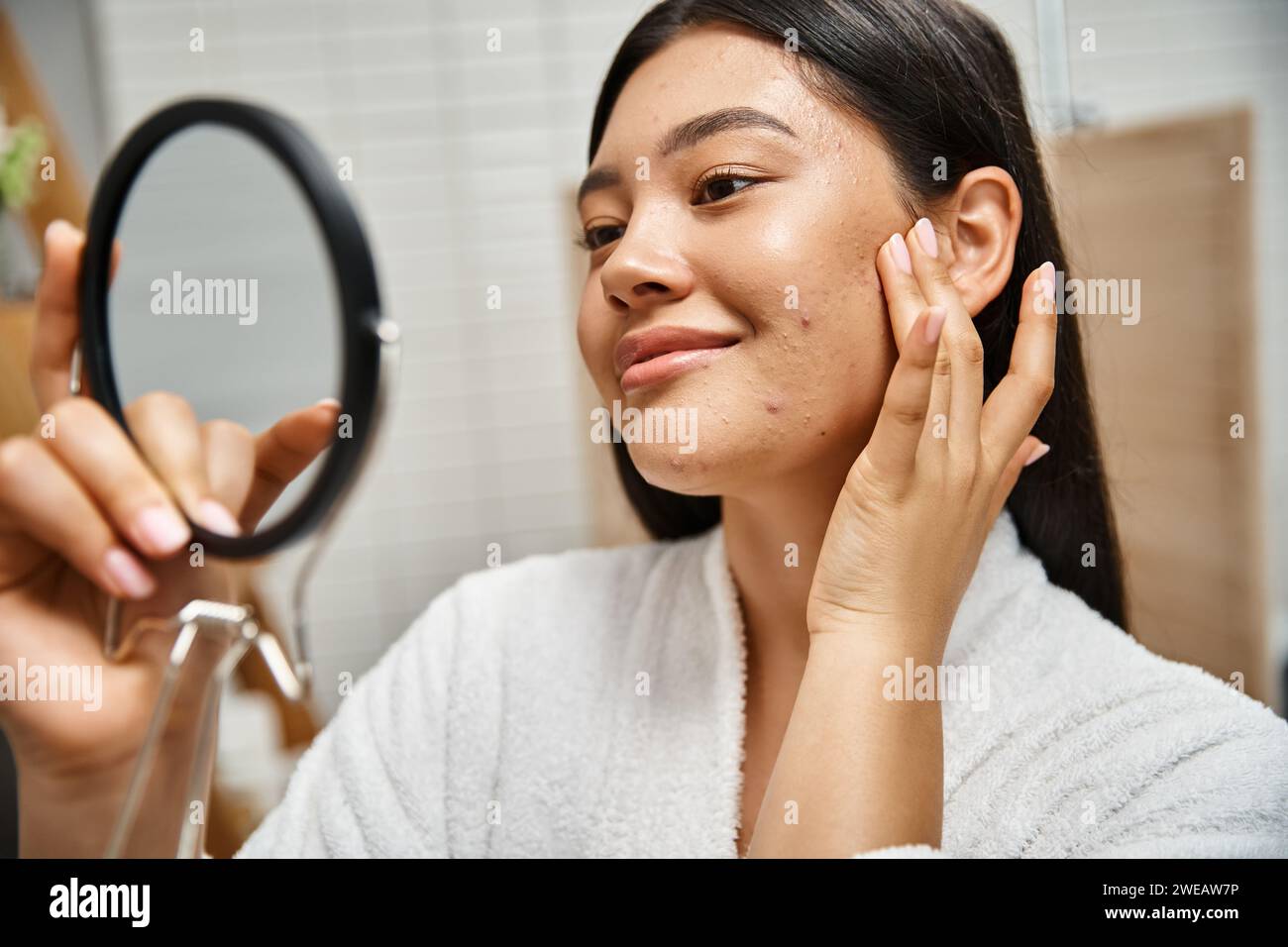 Junge und brünette asiatische Frau mit Pickel, die ihr Gesicht im Spiegel untersucht, Hautprobleme von echten Menschen Stockfoto
