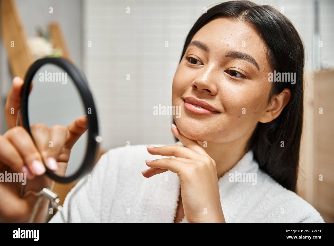 Junge und lächelnde asiatische Frau mit Pickel, die ihr Gesicht im Spiegel untersucht, Hautprobleme von echten Menschen Stockfoto