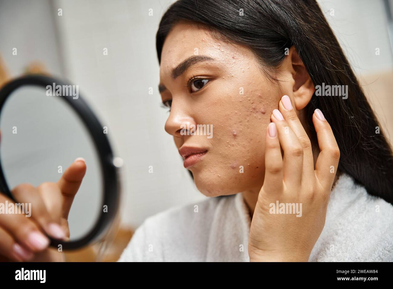Junge asiatische Frau mit brünetten Haaren und Pickel, die ihr Gesicht im Spiegel betrachtet, Hautprobleme Stockfoto