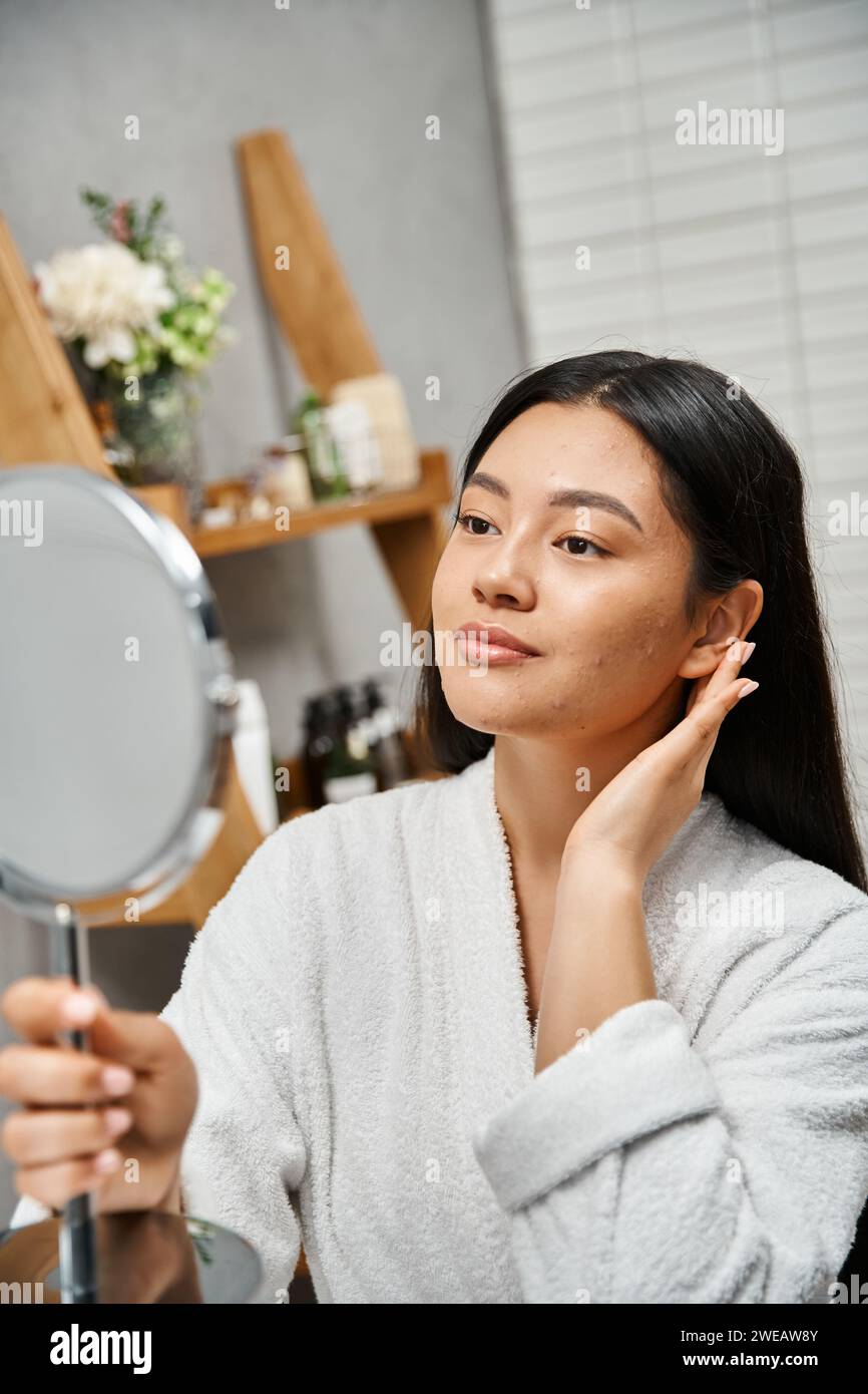 Porträt einer jungen asiatischen Frau im Gewand, die zu Akne neigende Haut berührt und im Badezimmer Spiegel betrachtet Stockfoto