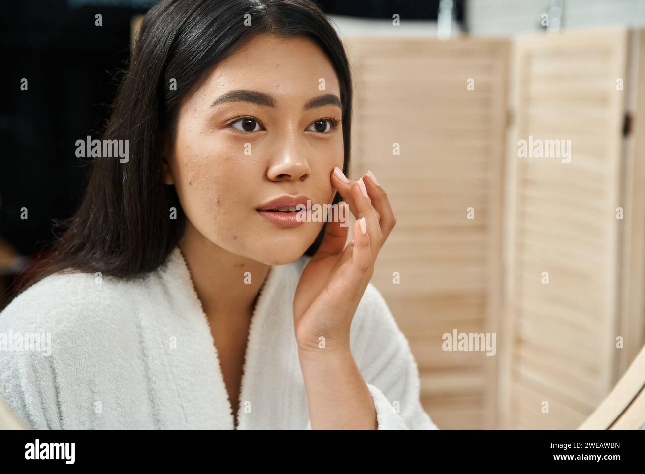 Junge asiatische Frau mit brünetten Haaren und zu Akne neigender Haut, die in den Spiegel im Badezimmer blickt, Hautpflege Stockfoto