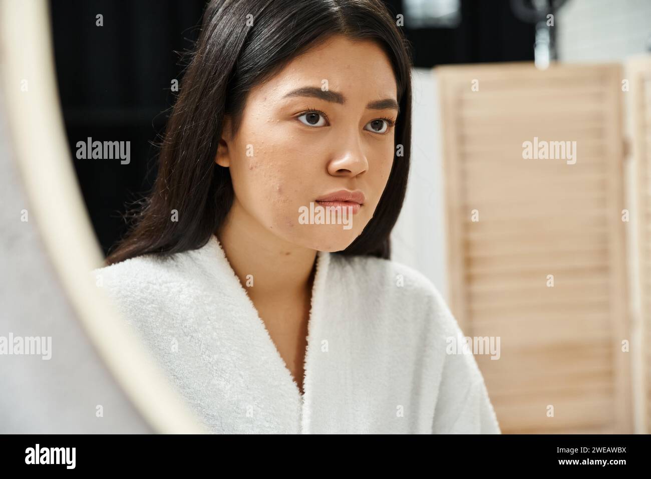 Traurige asiatische Frau mit brünetten Haaren, die ihr Gesicht mit Akne im Badezimmerspiegel untersucht, Hautproblem Stockfoto
