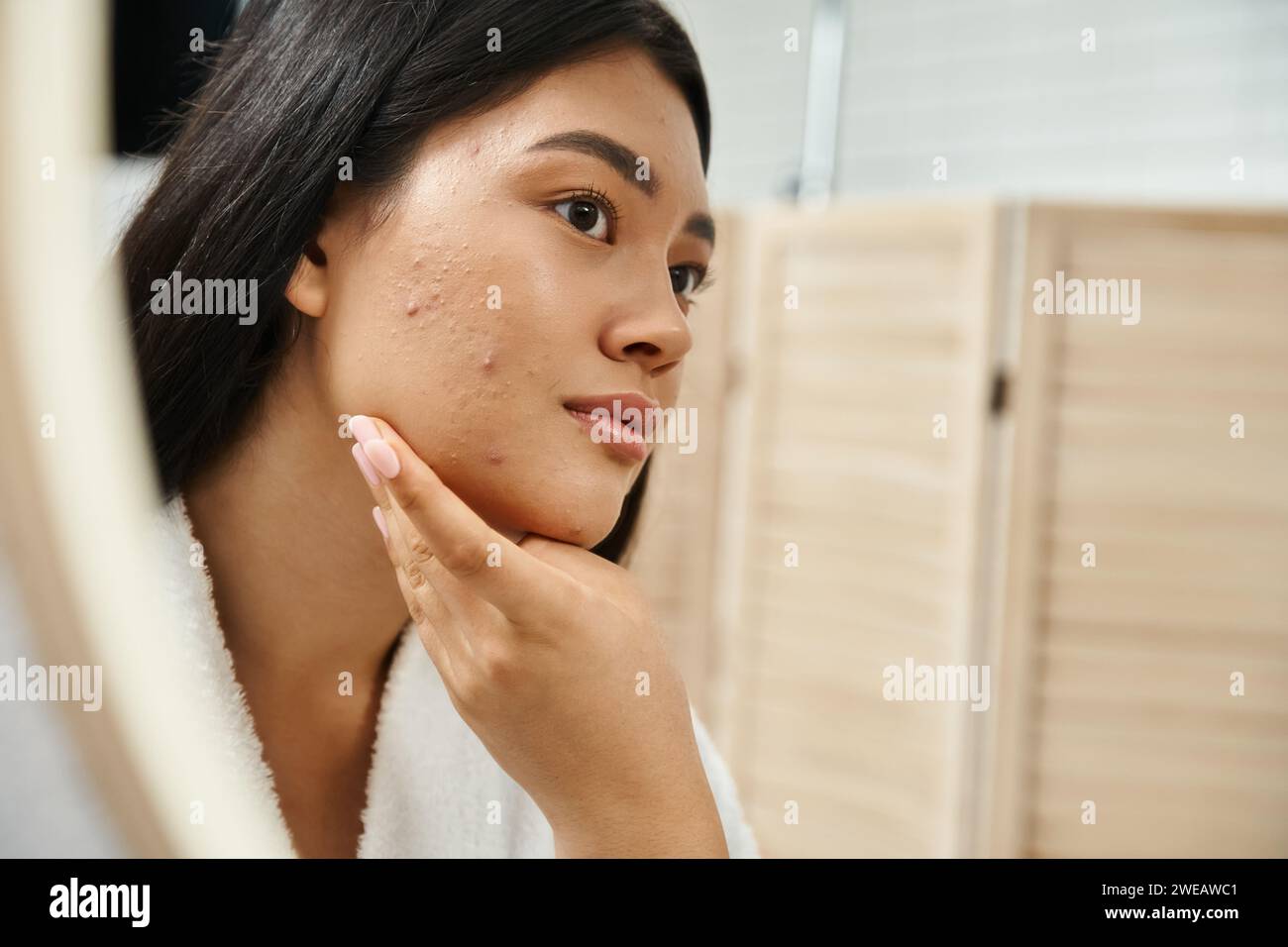 Junge asiatische Frau mit brünetten Haaren und Makel im Gesicht, Blick auf Spiegel im Badezimmer, Hautpflege Stockfoto