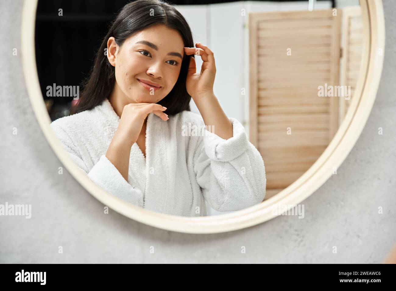 Fröhliches asiatisches Mädchen mit brünetten Haaren, das ihr Gesicht mit Akne im Badezimmerspiegel untersucht, Hautproblem Stockfoto