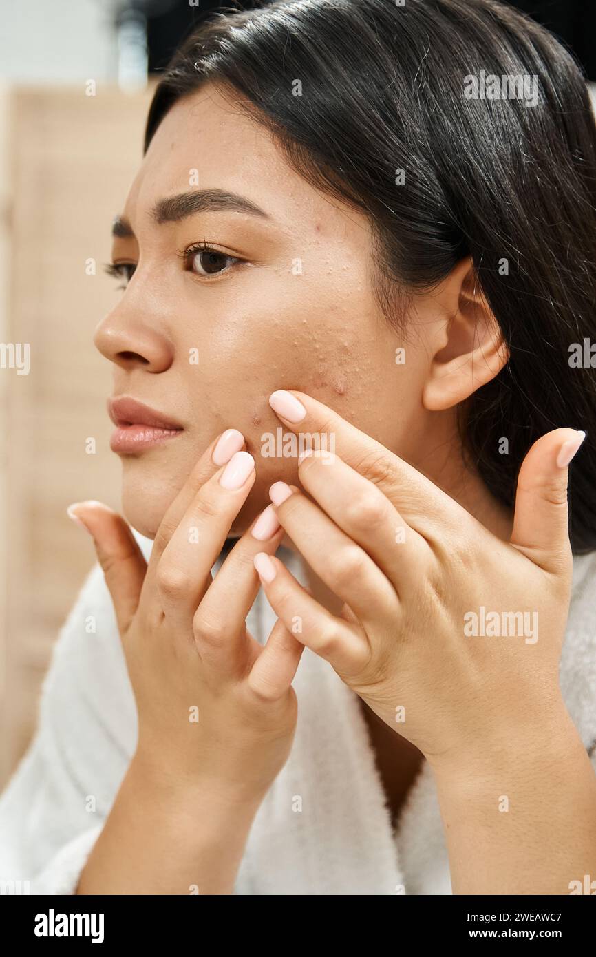 Junge asiatische Frau mit brünetten Haaren untersucht ihre Haut mit Akne im Badezimmerspiegel, vertikal Stockfoto