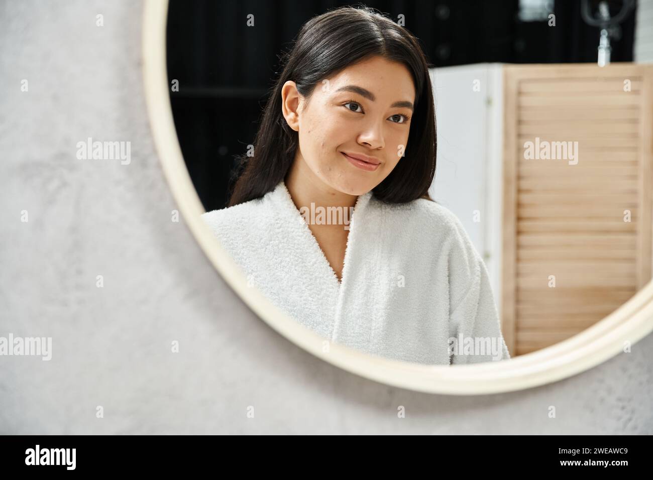 Glückliches asiatisches Mädchen mit brünetten Haaren, das ihr Gesicht mit Akne im Badezimmerspiegel untersucht, Hautproblem Stockfoto