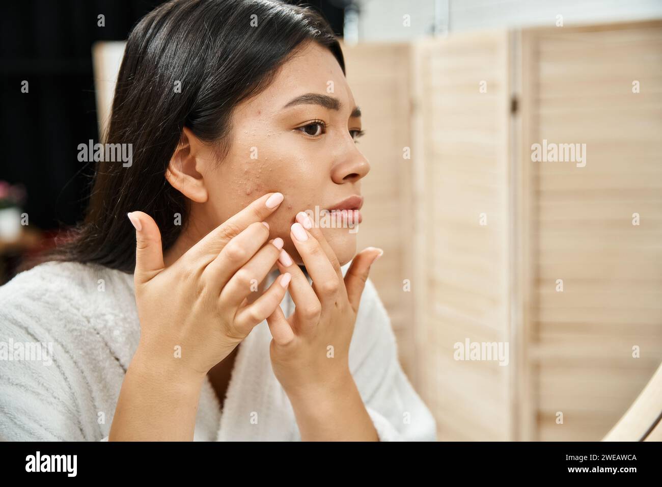 Junge asiatische Frau mit brünetten Haaren untersucht ihre Haut mit Akne im Badezimmerspiegel, Hautproblem Stockfoto