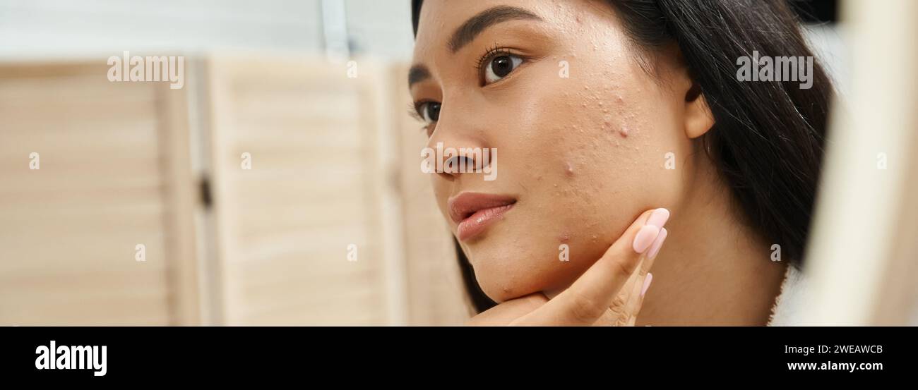 Junge asiatische Frau mit brünetten Haaren und zu Akne neigender Haut, die in den Spiegel im Badezimmer schaut, Banner Stockfoto