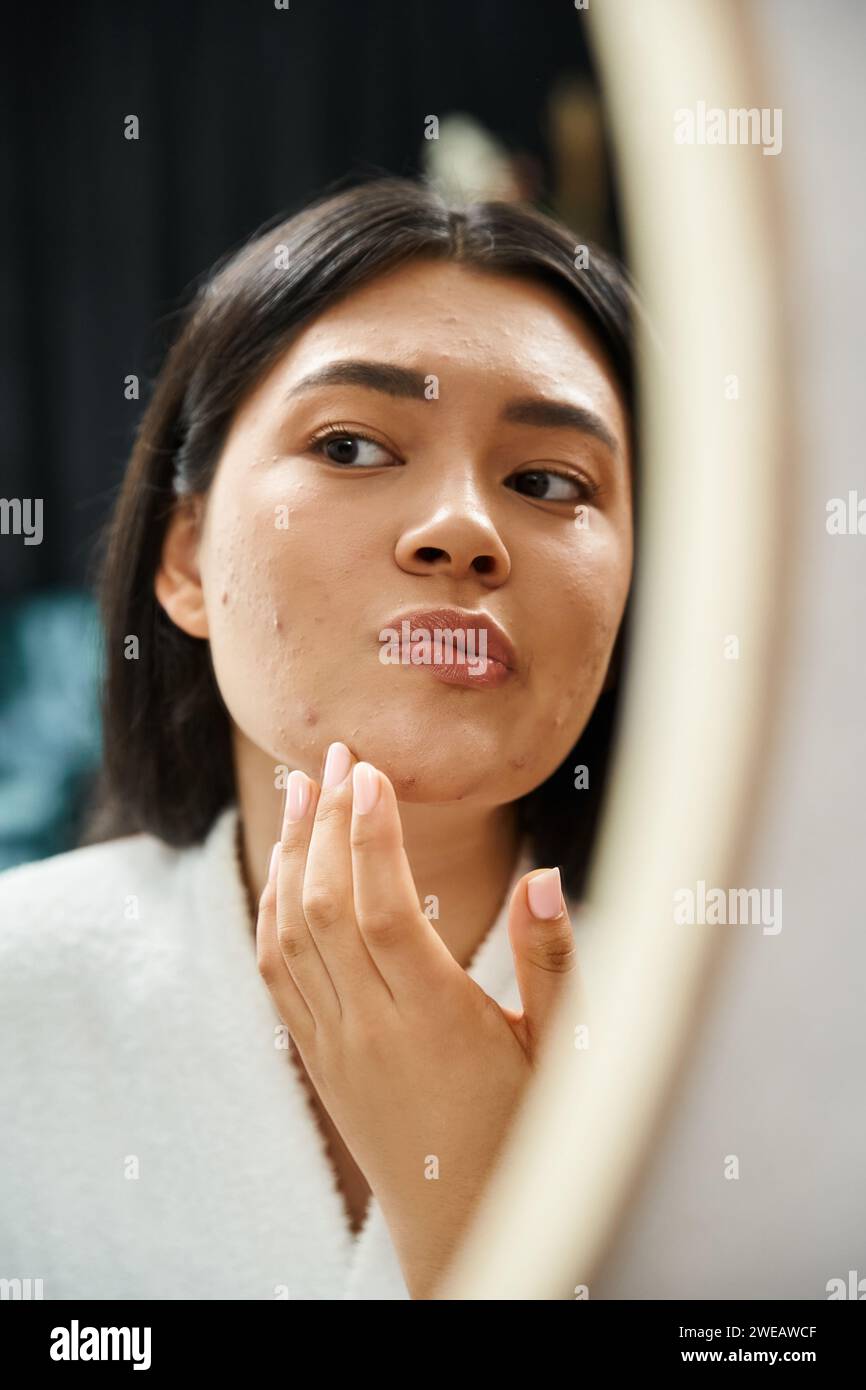 Junges asiatisches Mädchen mit brünetten Haaren, das ihr Gesicht mit Akne im Badezimmerspiegel untersucht, Hautproblem Stockfoto
