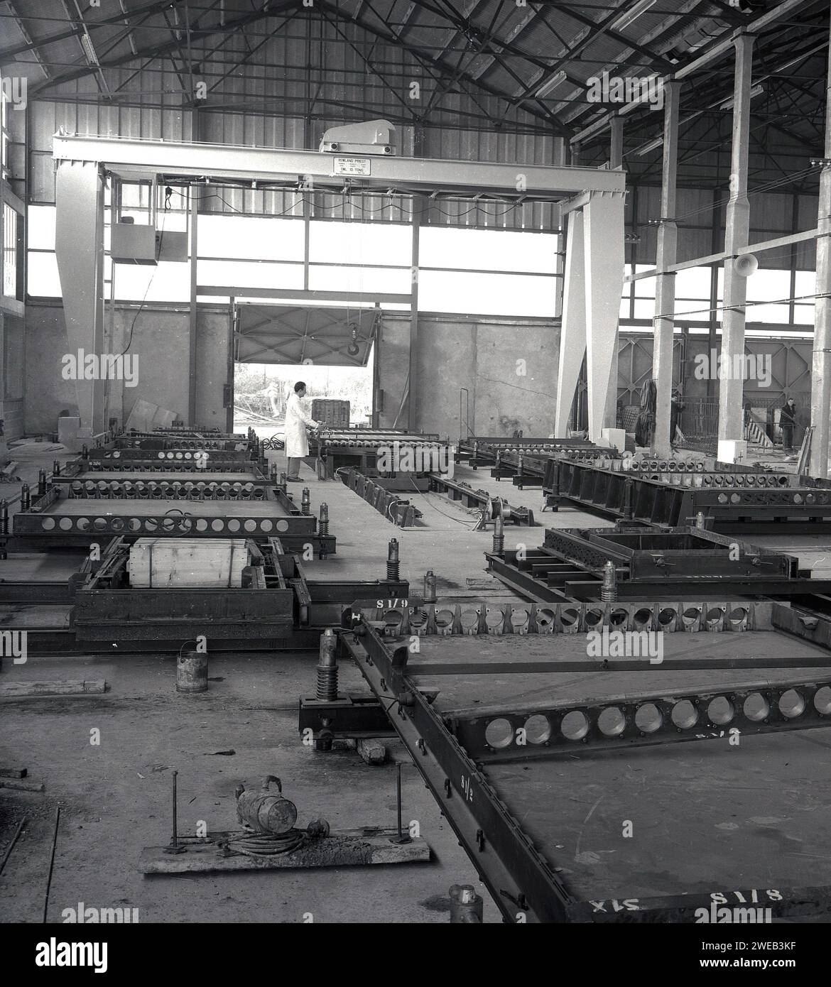 1960er Jahre, historisches Inneres eines großen „Hangar“-Gebäudes mit Grantry, in dem Fertigbetonplatten hergestellt werden, England, Großbritannien. Name oben auf der Gantry, Rowlan Priest, Cradley Heath, SWL 10 Tonnen. In Großbritannien in den späten 50er und 60er Jahren wurden große Turm- und Büroblöcke aus vorgegossenen Betonplatten errichtet, was eine schnellere und kostengünstigere Konstruktion und eine hervorragende Feuerbeständigkeit bedeutet. Stockfoto