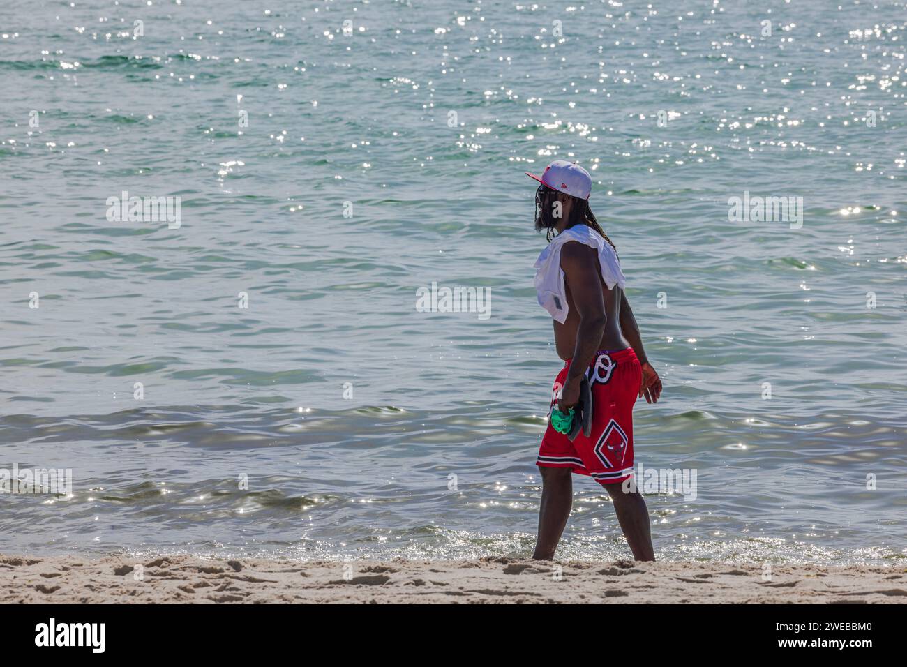 Ein hübscher junger Afroamerikaner mit sportlichem Aufbau, in roten Badeshorts und einer weißen Mütze, schlendert entlang der Küste von Miami Beach. USA. Stockfoto