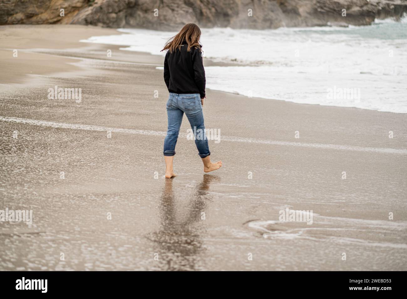 Erwachsene Frau, die im Winter entspannt an einem Strand spaziert und die Auswirkungen des Wassers beobachtet, das am Ufer fließt. Rückansicht in voller Länge, Kopierraum. Stockfoto