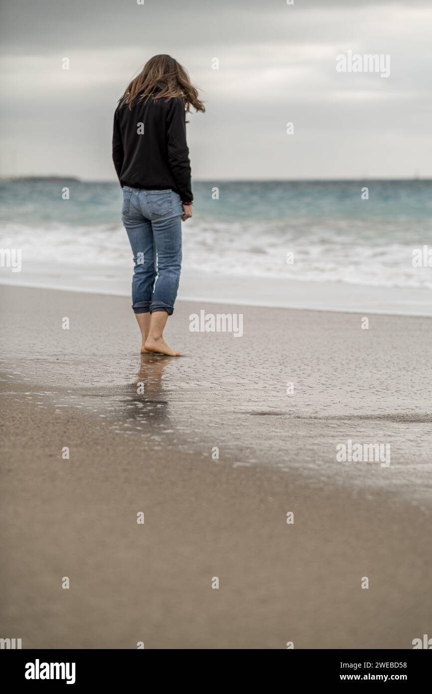 Die volle Rückansicht einer langhaarigen Frau, die barfuß am Wasserrand steht. Vertikale Komposition, Kopierraum. Stockfoto