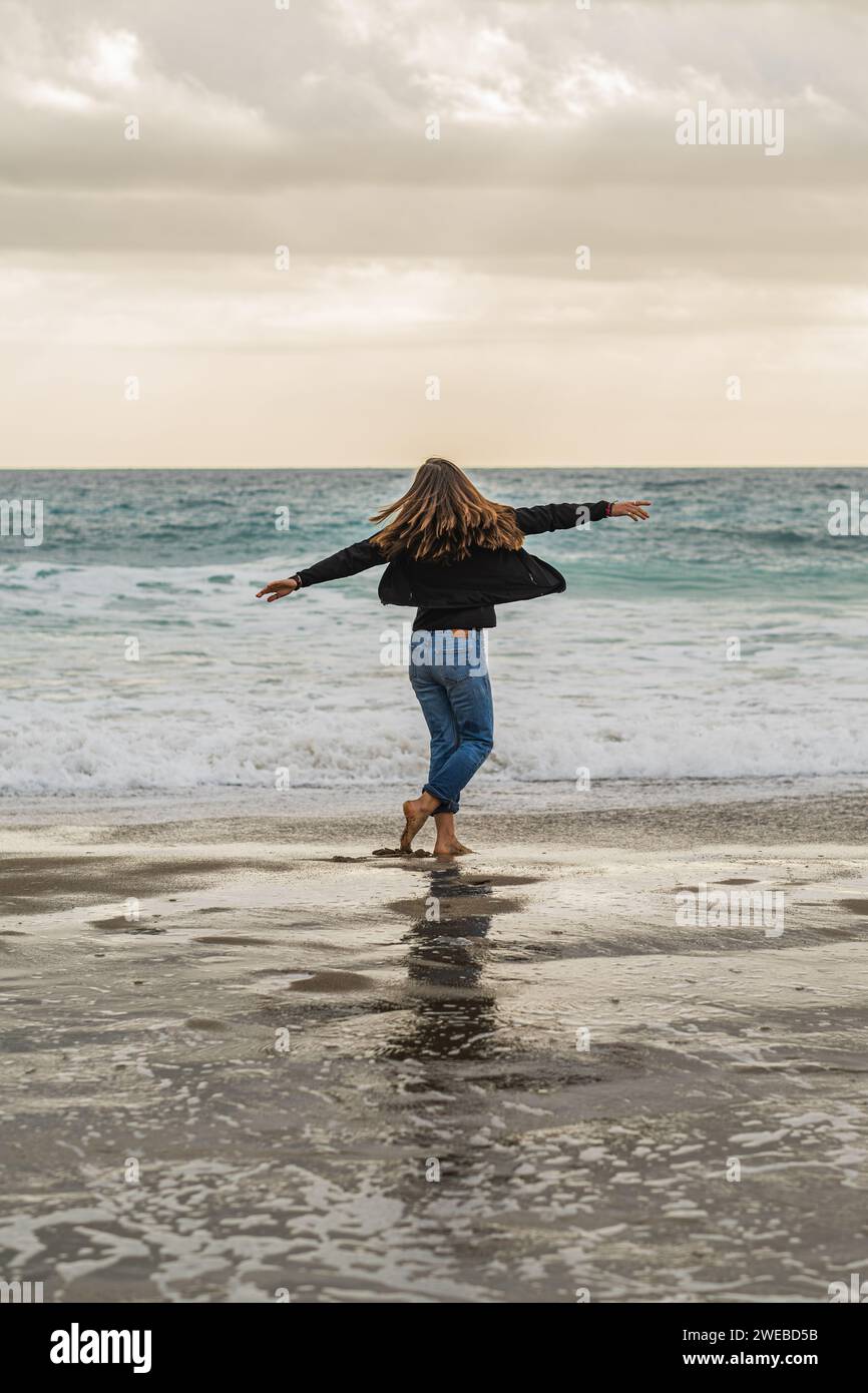 Frau in Bewegung, tanzt glücklich am Strand, Arme breit und lange Haare im Wind, Rückansicht. Kostenloses und fröhliches Lifestyle-Konzept. Stockfoto