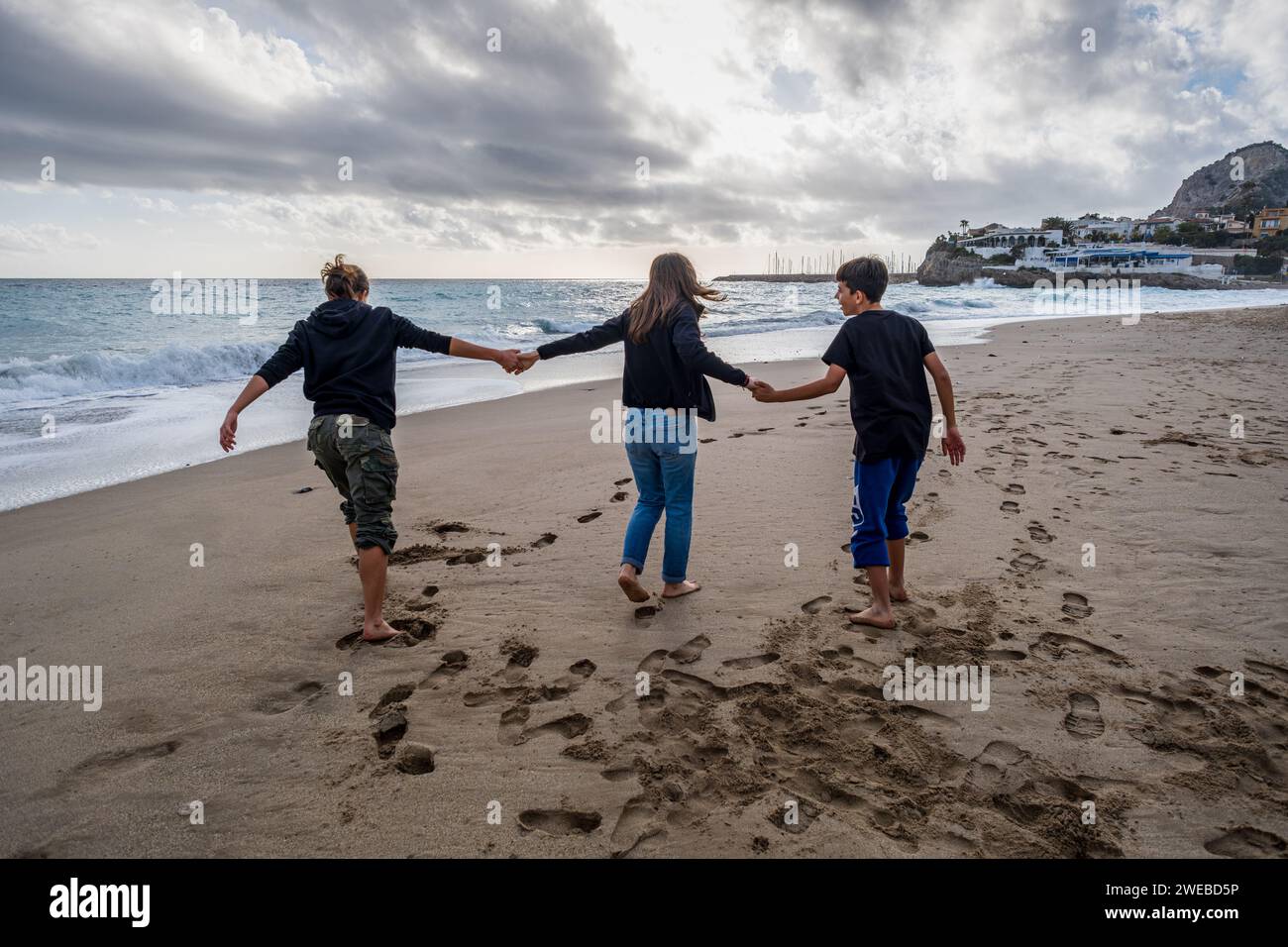 Rückansicht einer jungen Mutter und ihrer Söhne im Teenageralter, die an einem einsamen Strand gemeinsam Zeit genießen, Hände halten und an einem bewölkten Tag spazieren gehen. Stockfoto