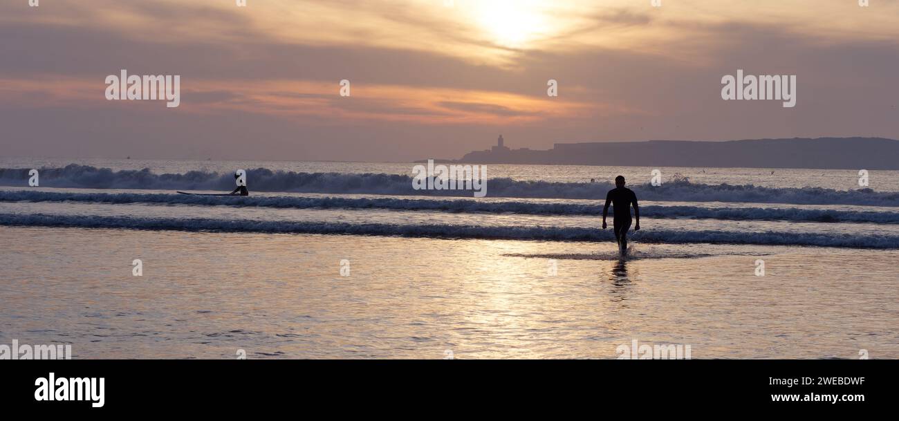 Ein Mann in Silhouette geht vom Wasser weg, während ein Surfer die Wellen bei Sonnenuntergang mit der Insel dahinter in Essaouira, Marokko, am 24. Januar 2024 angreift Stockfoto