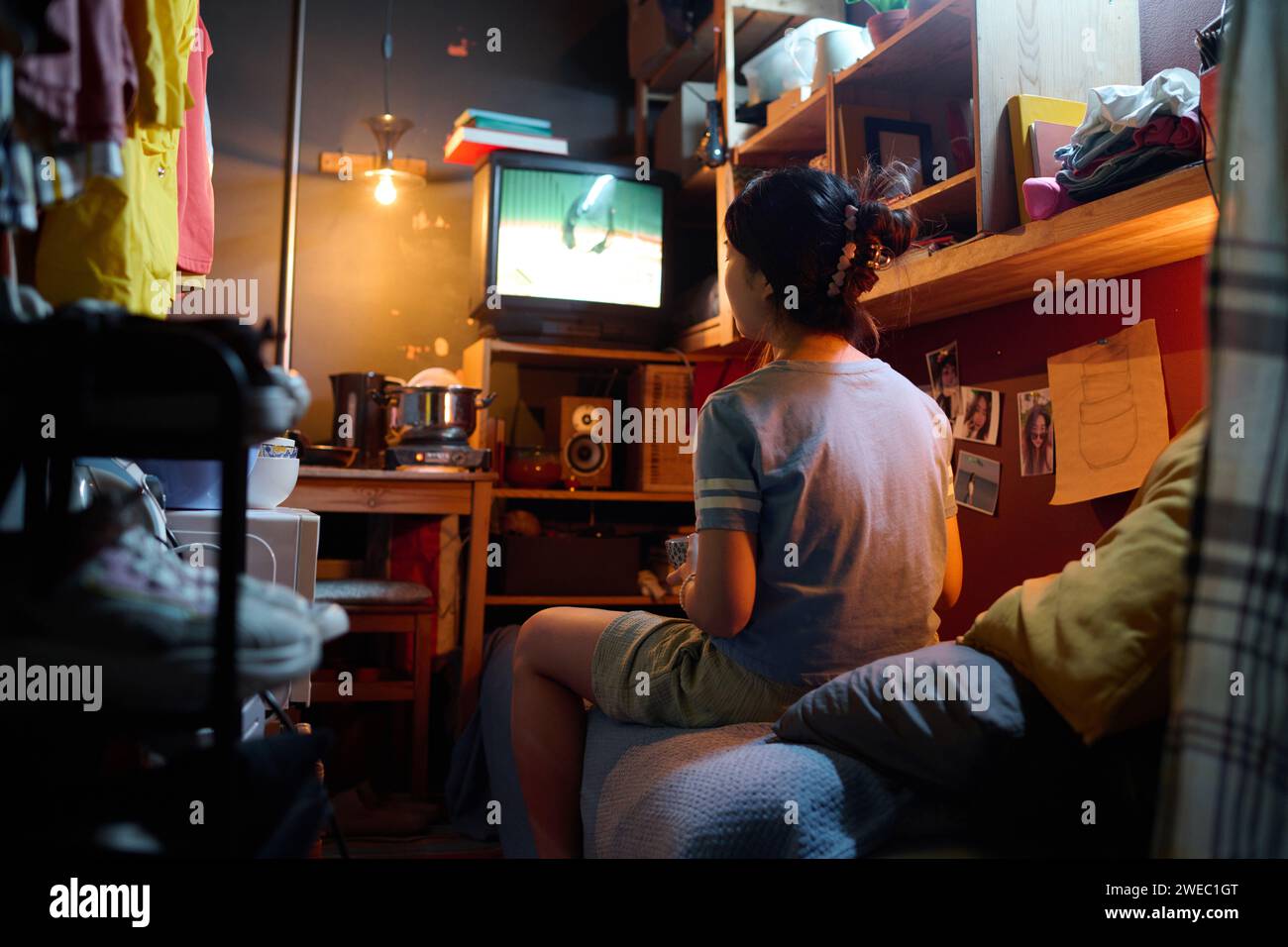 Rückansicht einer jungen asiatischen Frau in grauem T-Shirt, die auf dem Bett vor dem Bildschirm des Fernsehers sitzt und in Ruhe Fernsehen sieht Stockfoto