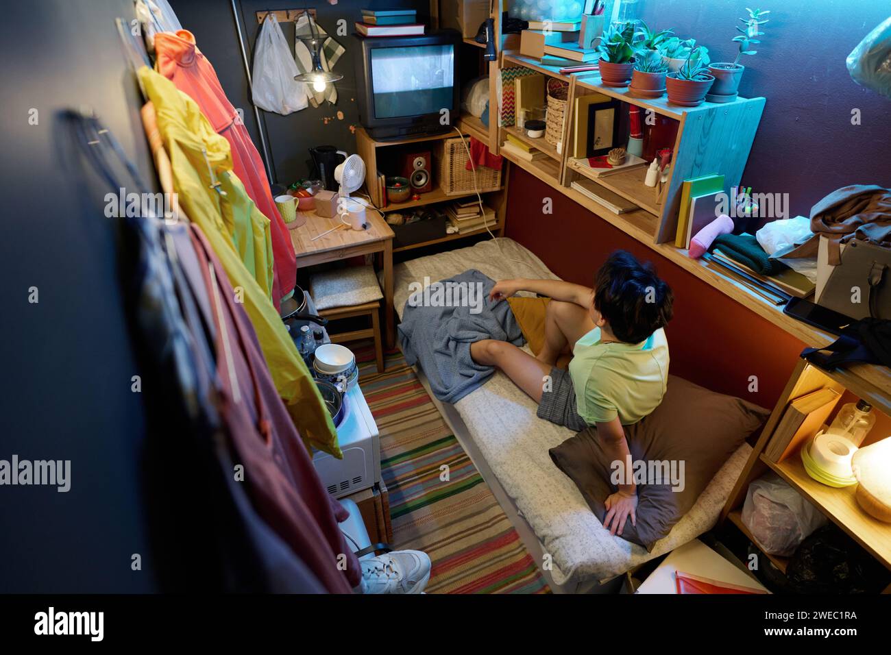 Hoher Winkel einer jungen asiatischen Frau in Casualwear, die auf einem Einzelbett vor dem Bildschirm des alten Fernsehers sitzt und Fernsehen oder Filme ansieht Stockfoto