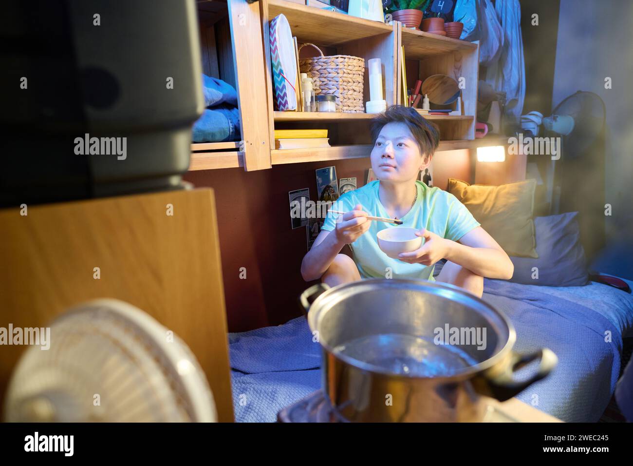 Asiatisches Mädchen mit Schüssel und Essstäbchen, das auf dem Bett vor dem Fernseher in einer kleinen Wohnung sitzt und beim Mittagessen Filme oder Sendungen ansieht Stockfoto