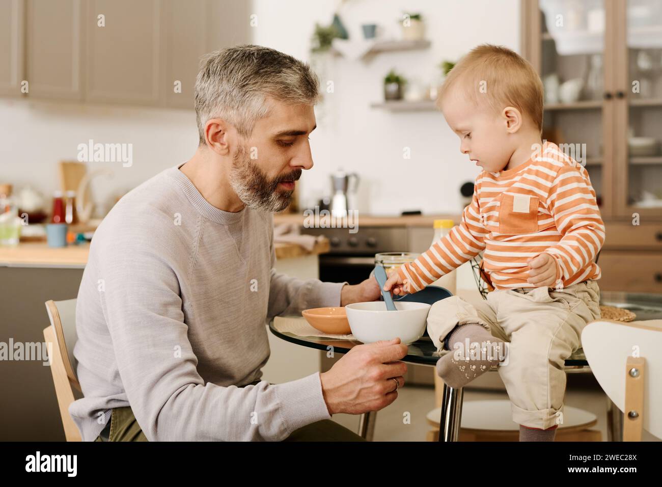 Der reife Vater sitzt am kleinen Tisch vor seinem liebenswerten Sohn und legt zum Frühstück einen Löffel in eine Schüssel mit Cornflakes oder anderen Speisen Stockfoto