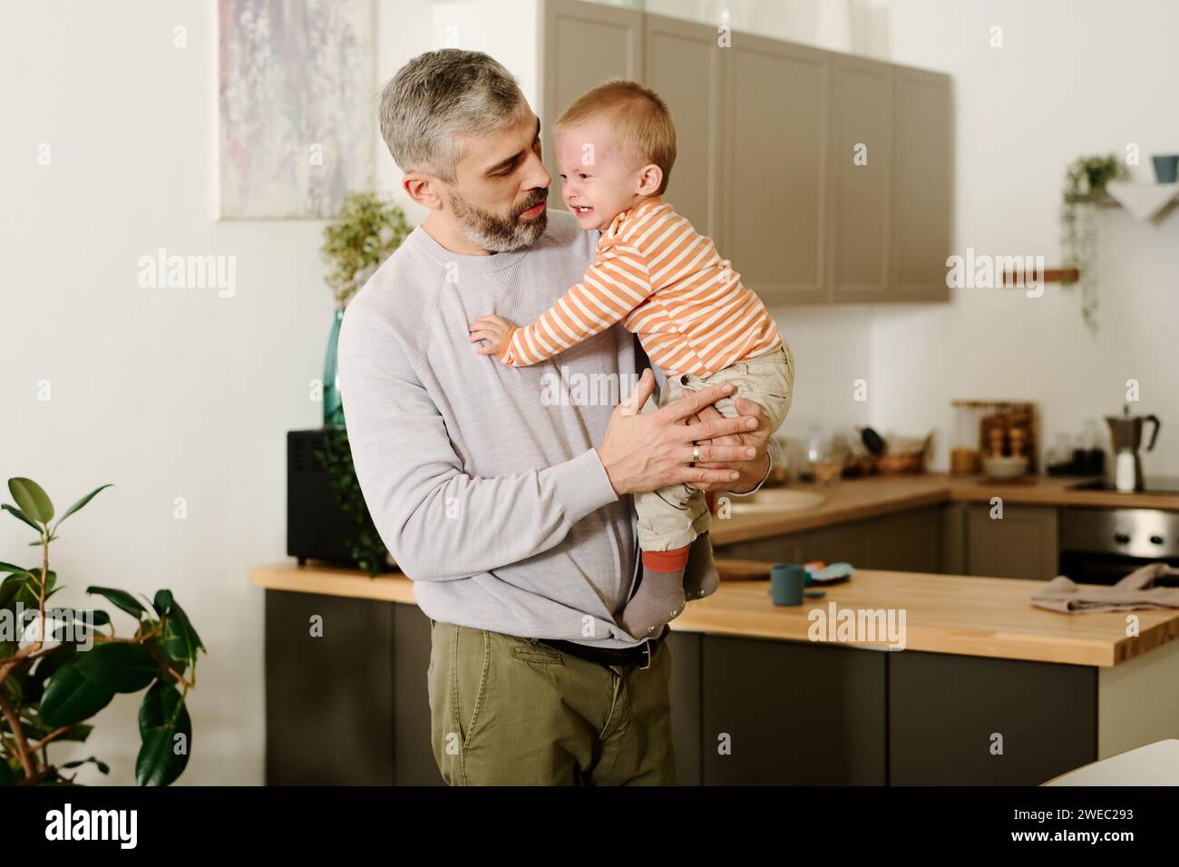 Reifer Mann in grauem Pullover und beigefarbener Hose hält seinen süßen frechen Sohn an den Händen und sieht ihn an, während er in der Küche steht Stockfoto